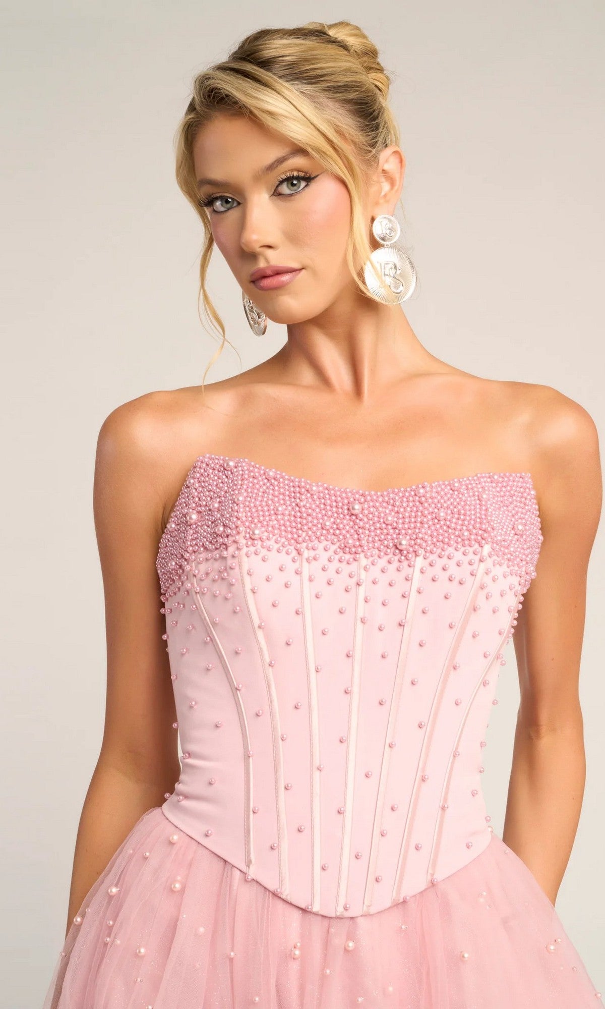 A woman poses against a neutral background wearing the Portia and Scarlett PS26034 long formal dress featuring a strapless corset bodice, glitter tulle skirt, bead embellishments, and large circular earrings.